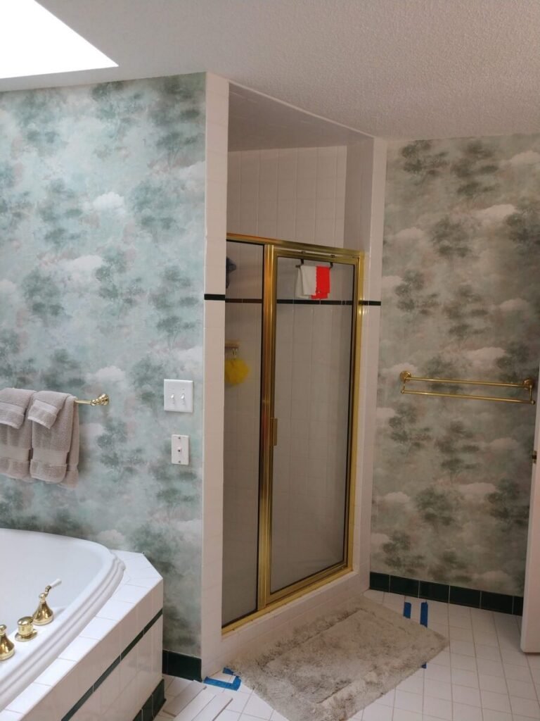 Dated master bathroom before remodel with brass shower door and cloud wallpaper