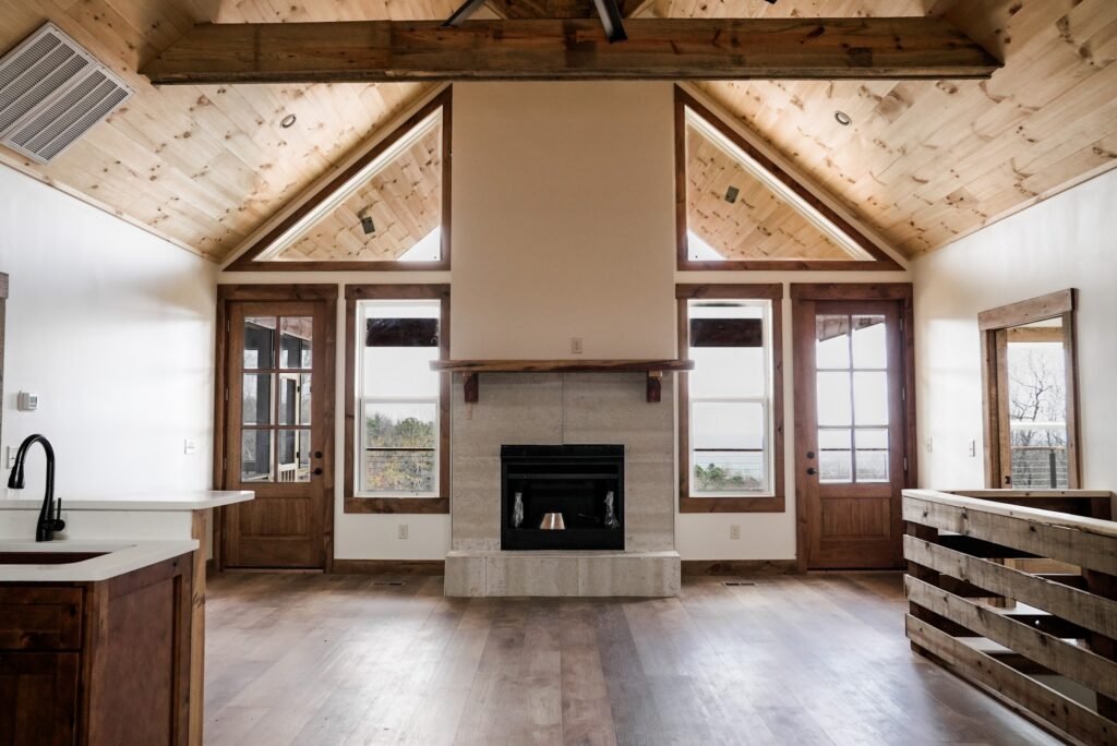 Open living area under a vaulted pine ceiling with exposed beams, gable windows, and a concrete-block fireplace with timber mantel