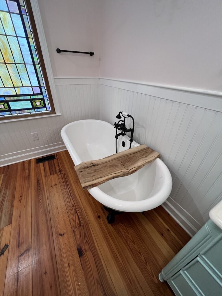 Angled view of white roll-top tub with live-edge wood caddy, black tub filler, beadboard walls and stained glass window