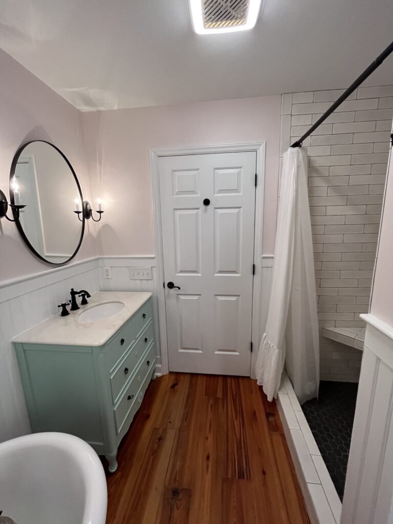 Full bath with mint green vanity, marble top, round black mirror, white subway tile shower and heart pine floors