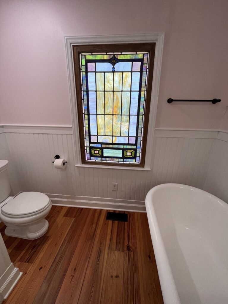 Tall leaded glass window in dark wood casing between a white toilet and the tub, over beadboard wainscot