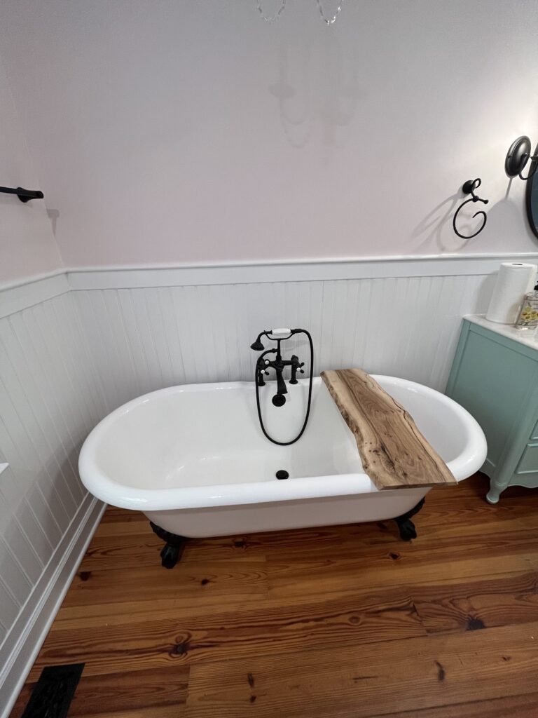 Freestanding tub on black claw feet with wooden bath board, black cross-handle filler and pale pink walls above beadboard