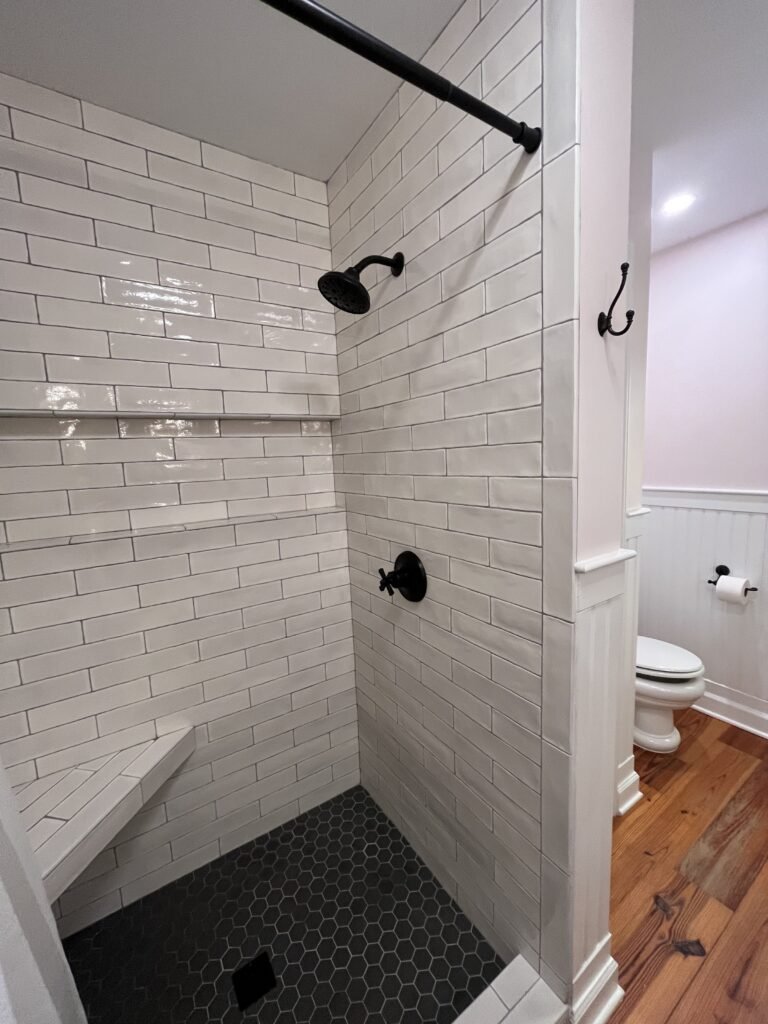 Walk-in shower lined in glossy white subway tile with corner bench, black fixtures and charcoal hexagon floor tile
