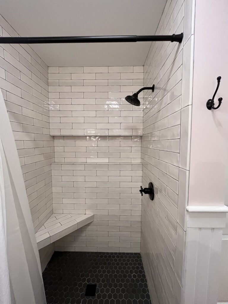 Straight-on shower alcove with tiled corner bench, black shower head and cross handle, and dark hexagon floor tile
