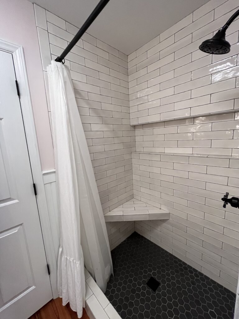 Shower seen from the doorway with white ruffled curtain drawn back, tiled bench seat and black hexagon floor
