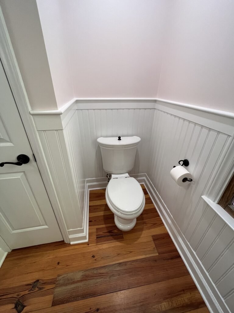 White two-piece toilet in a beadboard-paneled alcove with black paper holder, cap rail trim and warm pine flooring