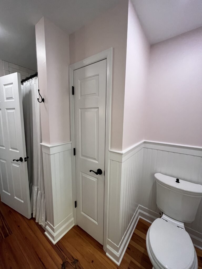 Blush pink walls above white beadboard wainscot, paneled closet door with black lever, shower curtain and toilet