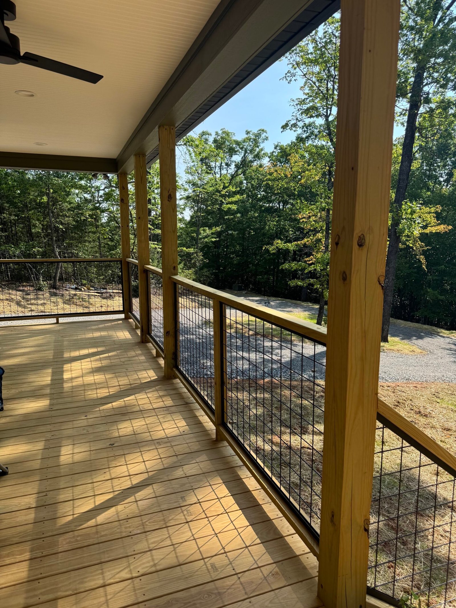Long covered porch looking down fresh wood decking past timber posts and wire railing toward gravel drive and forest