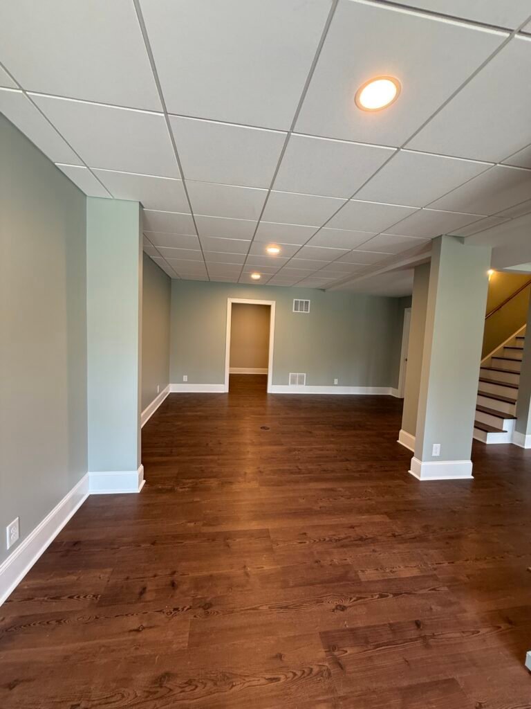 Open basement room in sage green with boxed support column, doorway to a back room, and staircase beyond