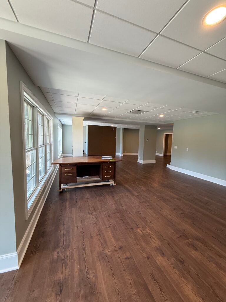 Daylight basement space with a bank of white-trimmed windows, dark wood desk, and open floor stretching toward distant rooms