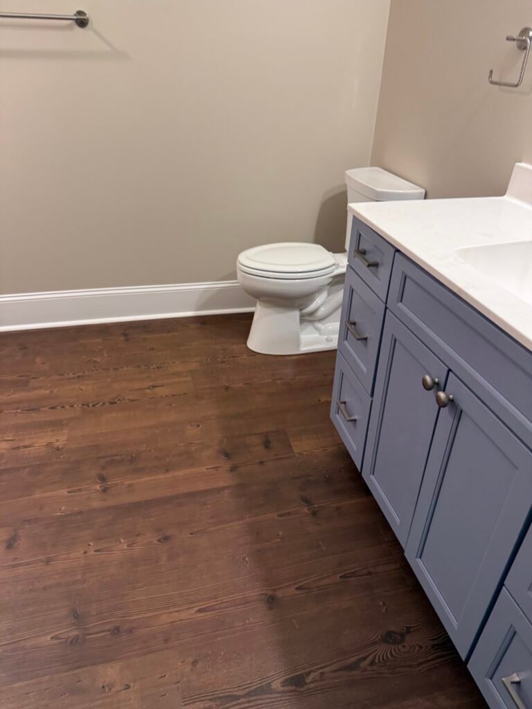 Basement bathroom with slate blue shaker vanity, white quartz-look counter, brushed nickel hardware, white toilet and dark plank floor
