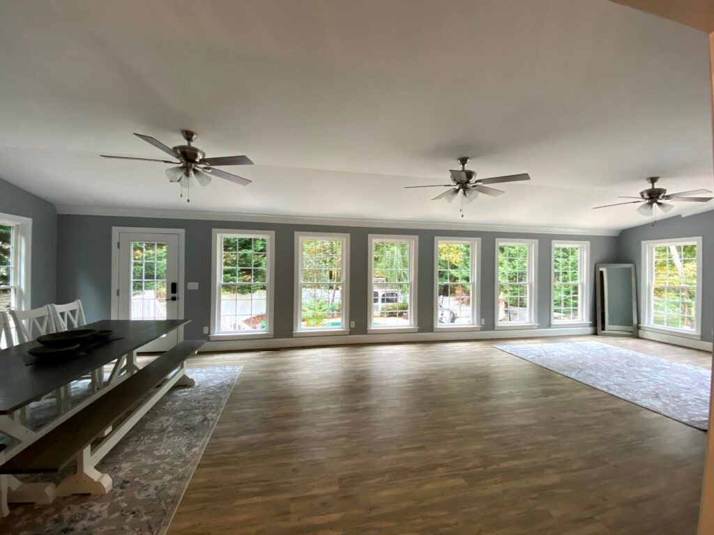 Wide open room with blue-grey walls, a row of white double-hung windows, three ceiling fans and plank flooring