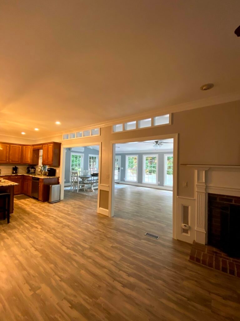 Brick fireplace with white mantel beside an oak kitchen, openings with transom windows revealing the sunlit blue addition beyond