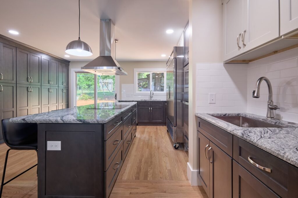 Galley view along veined grey granite island toward stainless refrigerator columns, with dark drawer bases and white subway tile