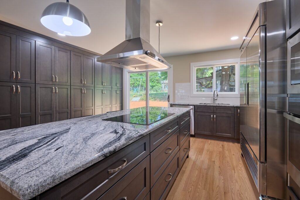 Dark stained cabinet wall beside granite island with black cooktop, stainless island hood, wall ovens and refrigerator at right
