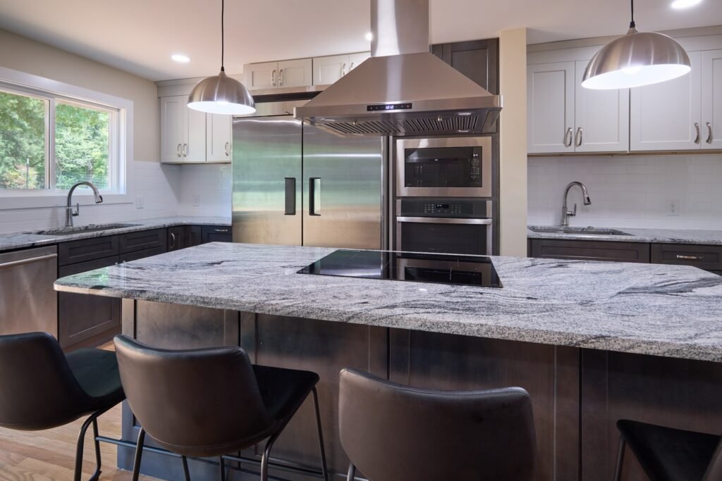 Granite island counter with black cooktop and leather barstools facing stainless refrigerator, built-in microwave and wall oven