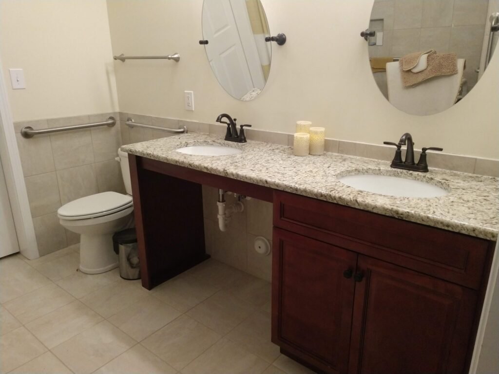 Roll-under double vanity with speckled granite top, dark cherry cabinetry, oval mirrors, and wall grab bars beside the toilet