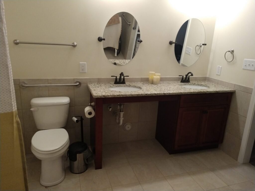 Wider view of the vanity wall: open knee space under one sink, grab bars flanking the toilet, tiled wainscot