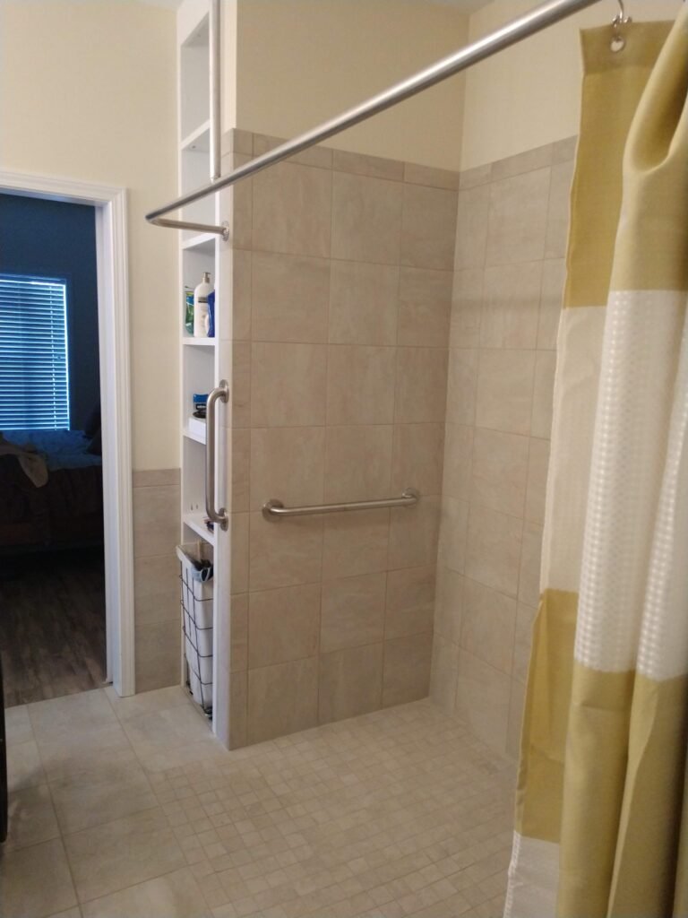 Half-wall shower partition with built-in shelving column and grab bar, level tile floor continuing toward the adjoining bedroom doorway