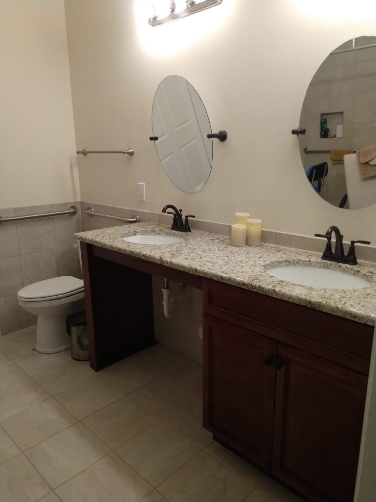 Angled view of the granite double vanity, brushed-nickel light bar, pivot mirrors, and open knee space beside the toilet