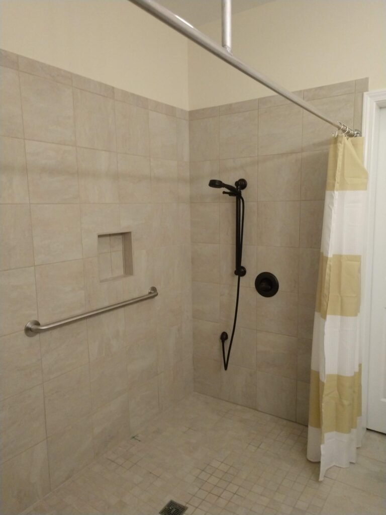 Corner of the walk-in shower showing tiled niche, long grab bar, bronze hand shower, and mosaic-tiled floor drain