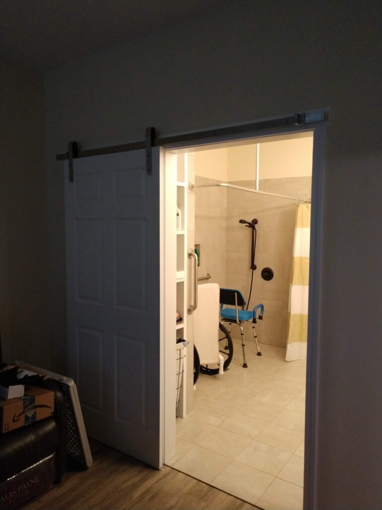 Sliding barn door rolled open on a black rail, revealing the tiled shower, shower chair, and wheelchair beyond