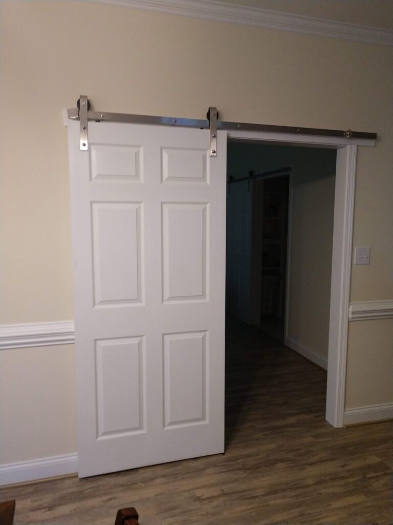 White six-panel barn door on a stainless track, slid open to a wide passage with wood-look plank flooring