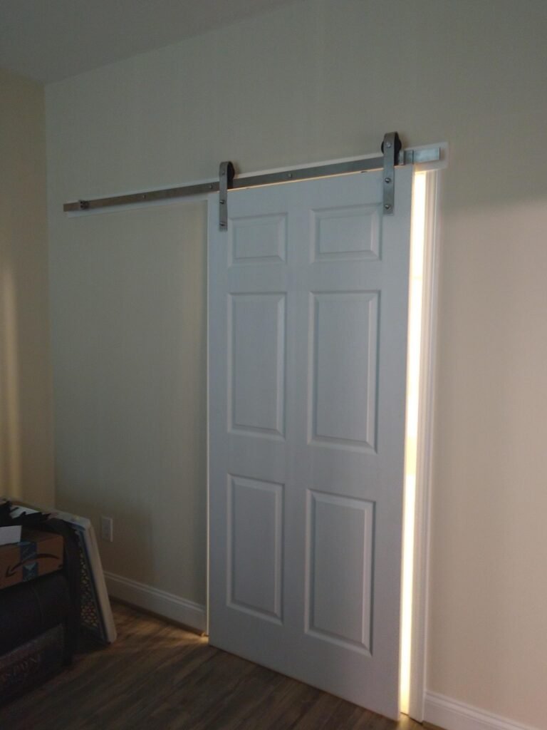 Closed white barn door on an exposed metal track against a pale wall, light glowing along the door's edge
