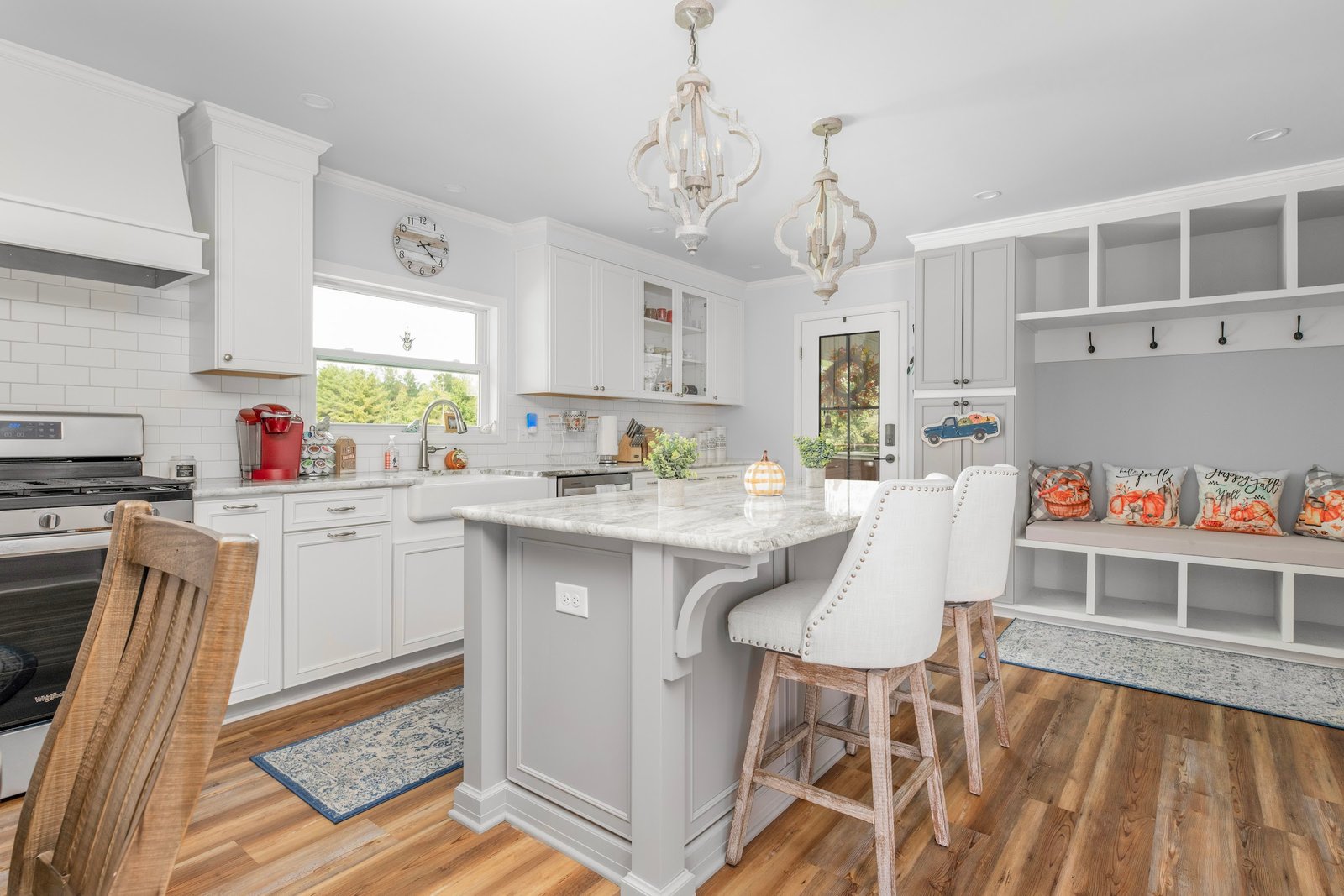 White shaker kitchen with grey marble-topped island, twin quatrefoil chandeliers, and a built-in mudroom bench beyond