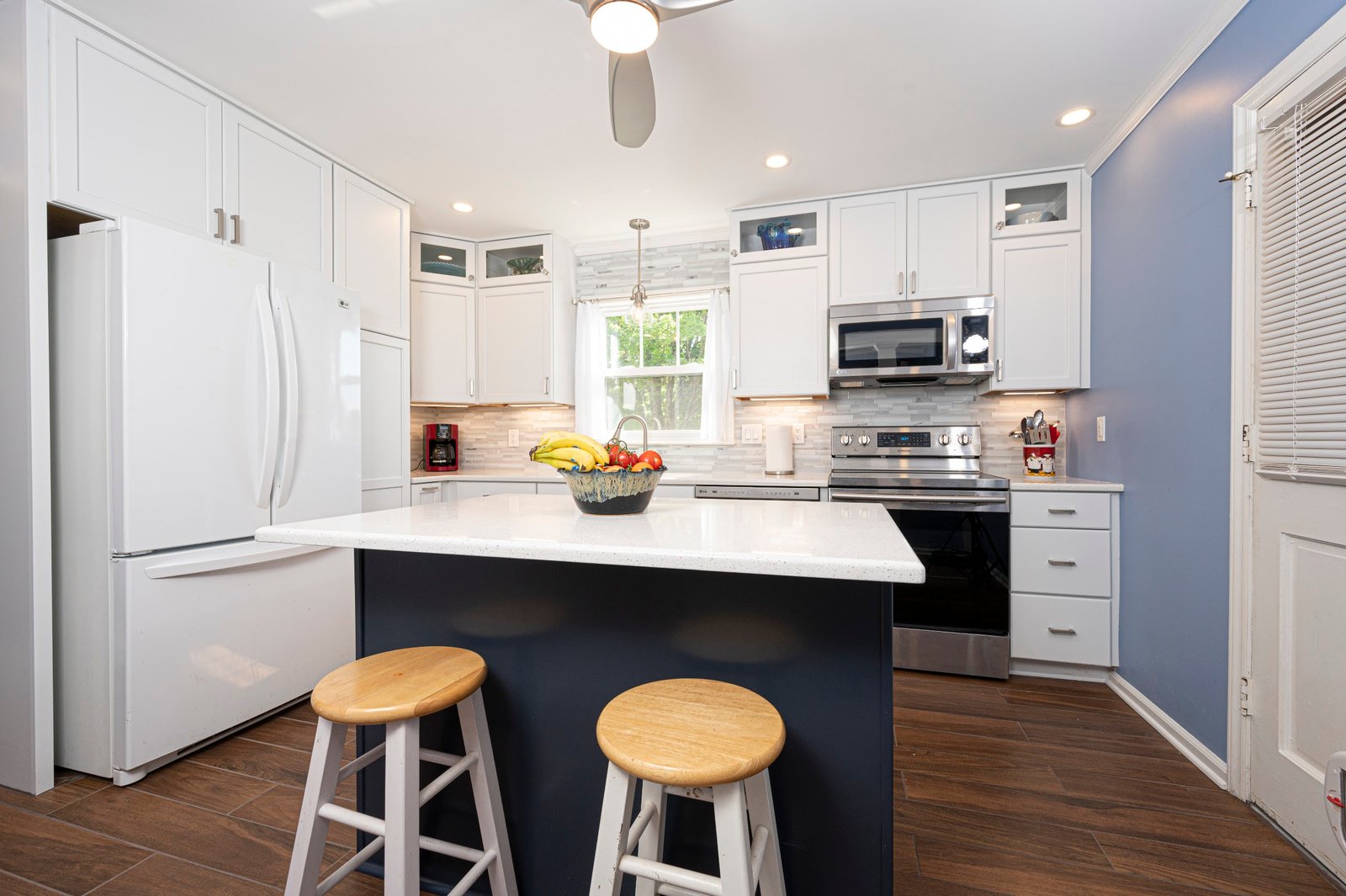 Wide kitchen view with navy quartz-topped island, two wood stools, white shaker cabinets and periwinkle wall